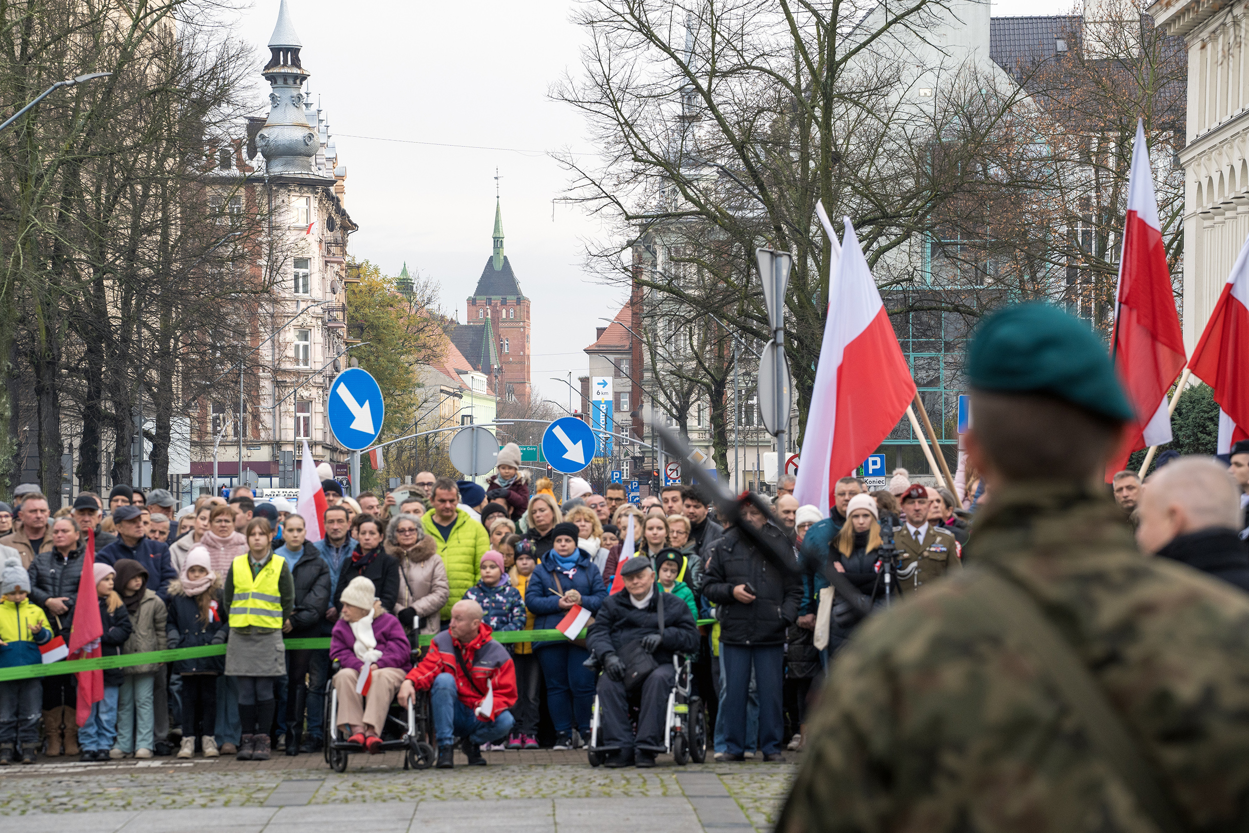 oficjalne uroczystości na pl. Józefa Piłsudzkiego w Gliwicach. Na pierwszym planie wojskowy stojący tyłem, na drugim planie mieszkańcy z biało-czerwonymi flagami. W tle budynki
