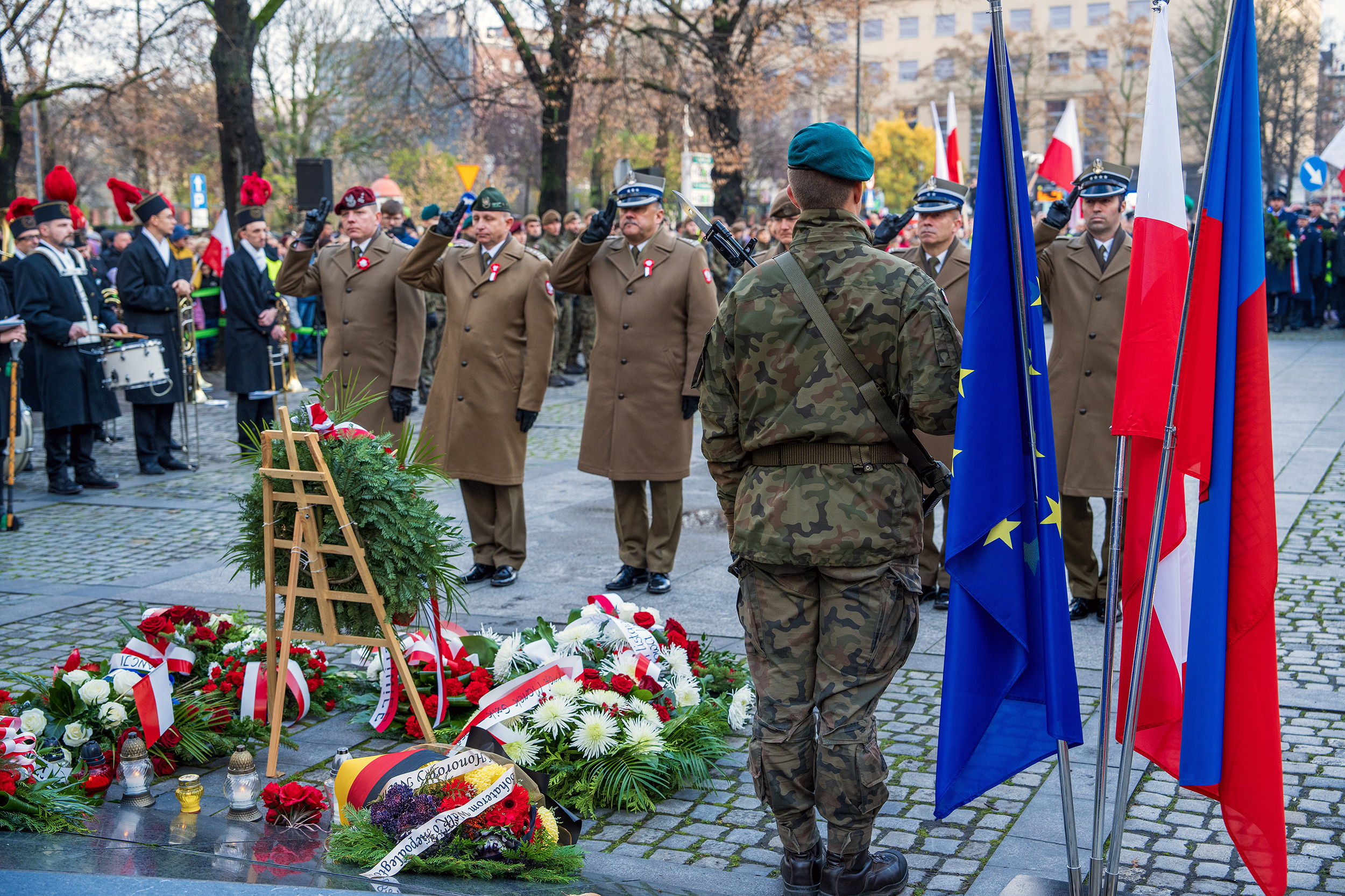 oficjalne uroczystości na pl. Józefa Piłsudzkiego w Gliwicach. Na pierwszym planie wojskowy stojący tyłem, na drugim planie wojskowi składający kwiaty pod pomnikiem. W tle orkiestra górnicza KWK Sośnica