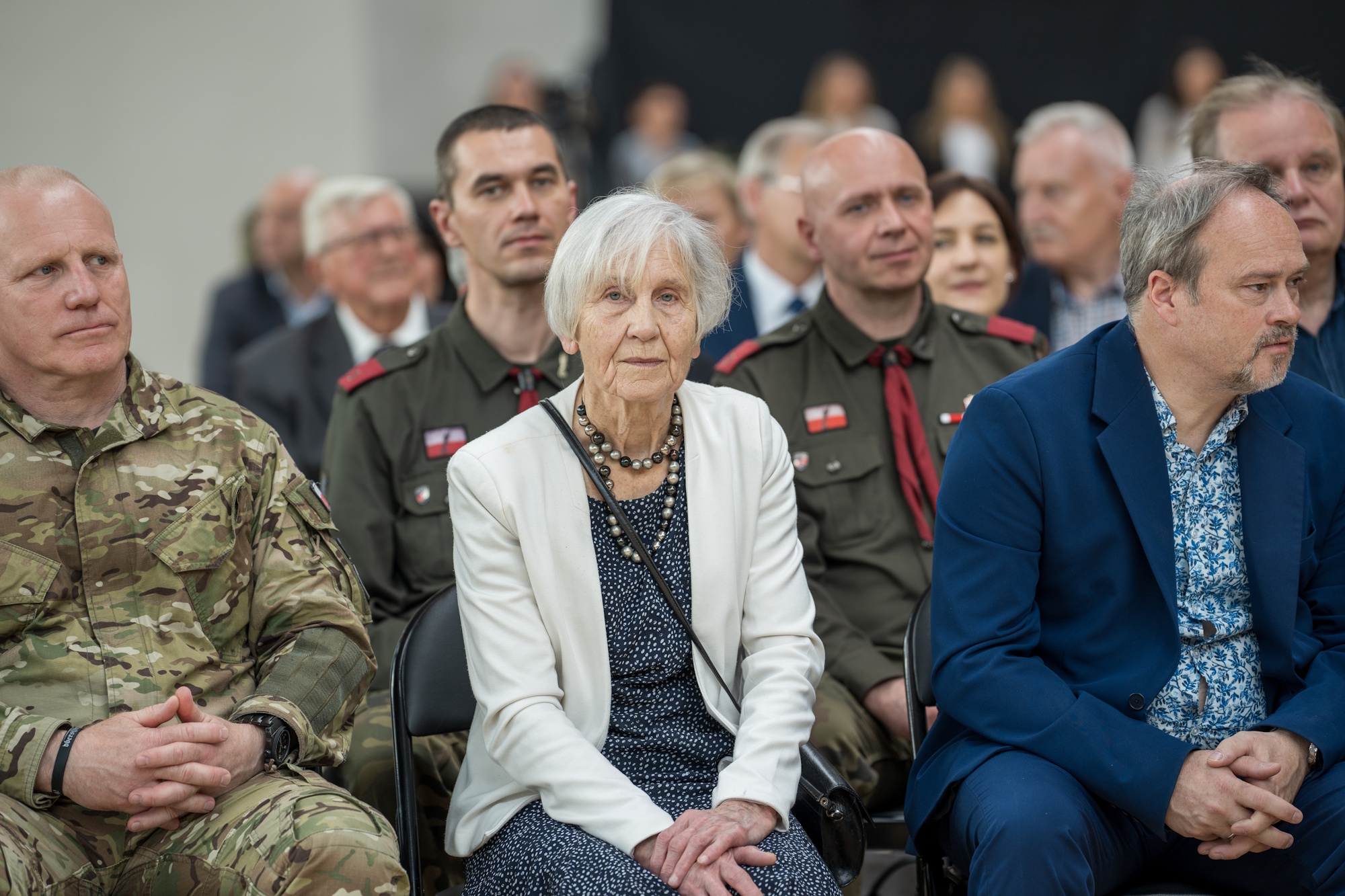 kobieta w białej marynarce siedzi w tłumie ludzi 