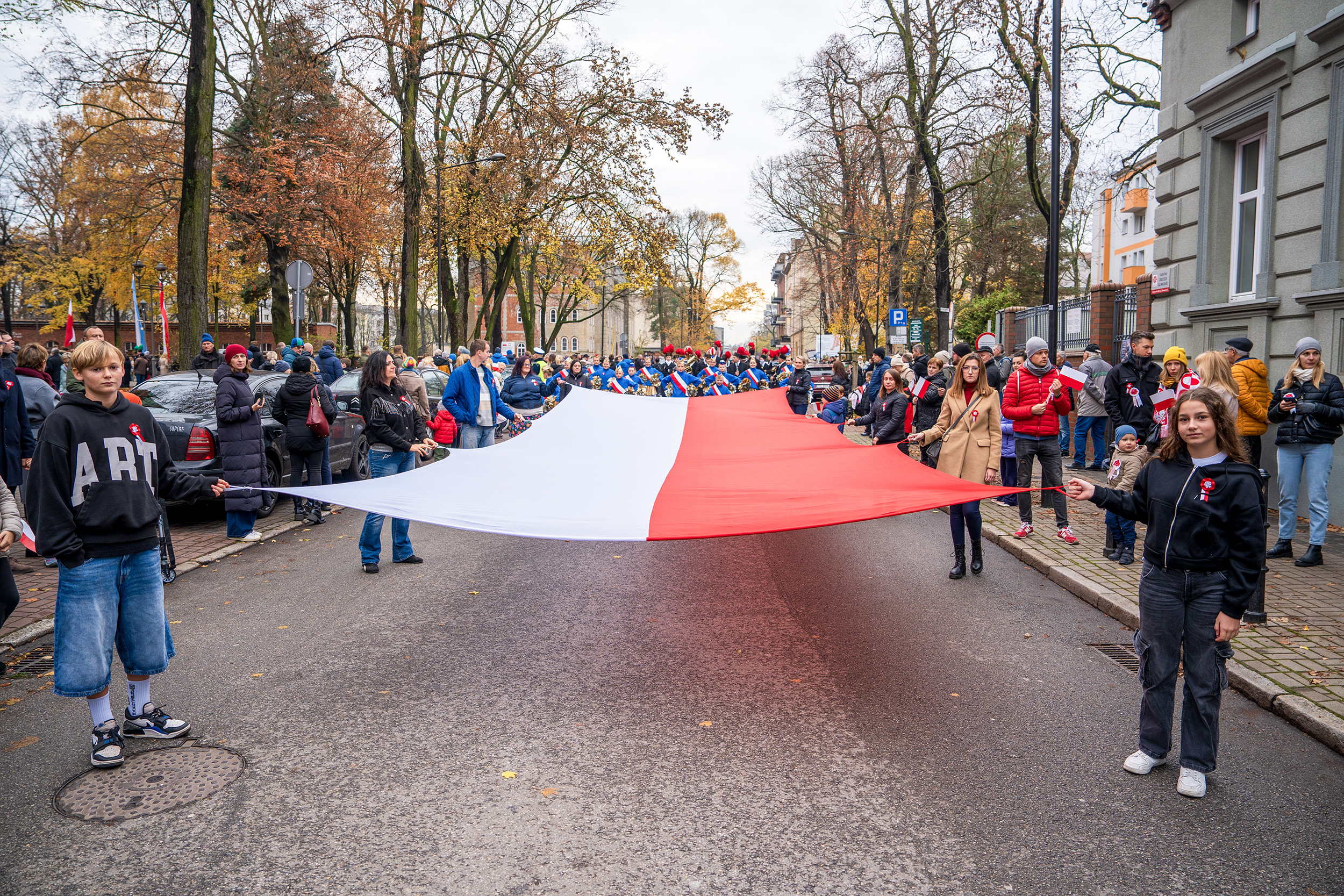 Przemarsz przez miasto z flagą Polski. Na pierwszym planie dzieci trzymające za rogi biało-czerwonego materiału
