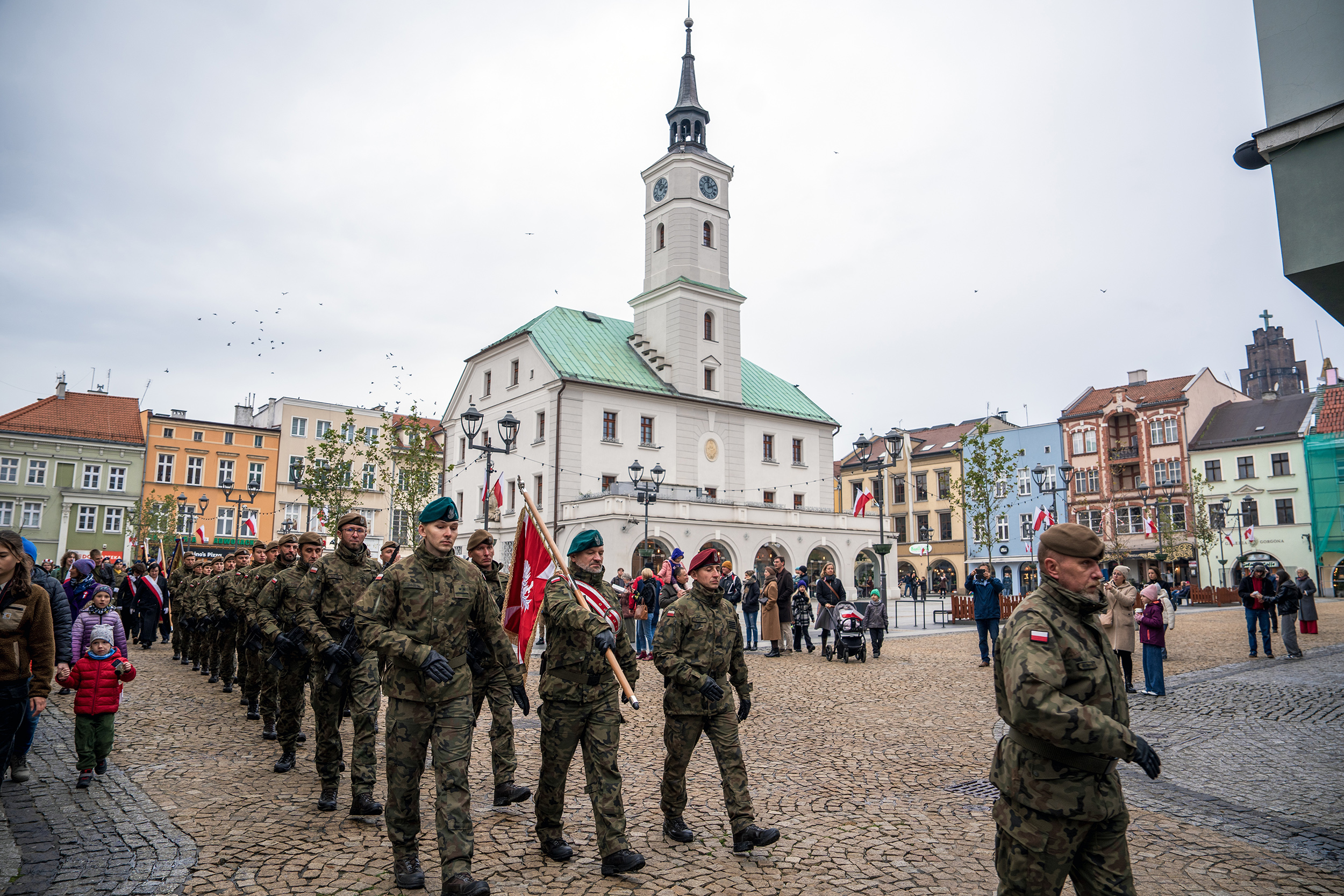 Przemarsz przez miasto z flagą Polski. Na pierwszym planie maszerujący wojskowi. W tle gliwicki Ratusz.
