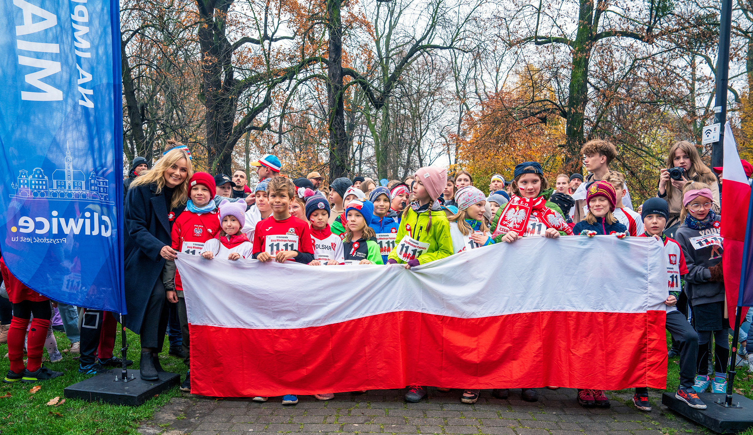 Dzieci przygotowane do Biegu Młodego Patrioty w parku Chopina za flagą Polski. Wśród dzieci prezydent Katarzyna Kuczyńska-Budka