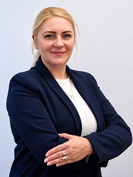 Kobieta o prostych blond włosach ubrana w granatową marynarkę i białą bluzkę